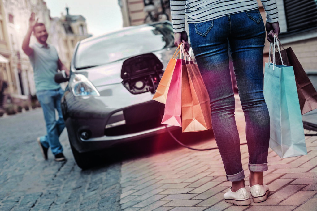 So many bargains. Selective focus on a young lady wearing casual attire carrying packages after having fun and buying many things returning to her cheerful boyfriend charging their car.