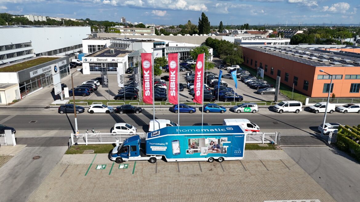 Homematic IP Truck von Siblik auf einem Parkplatz vor einem großen Bürokomplex, mit mehreren Fahrzeugen und Flaggen im Hintergrund.