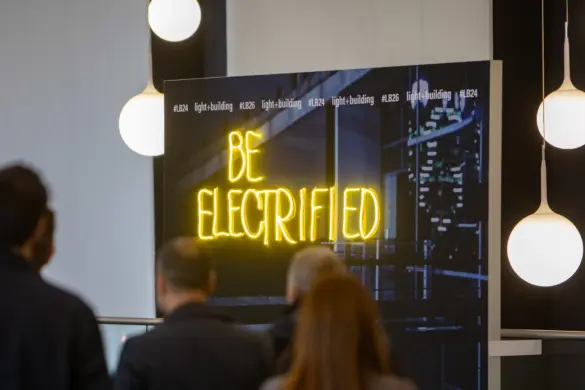 Besucher*innen gehen auf ein leuchtendes Schild mit der Aufschrift „BE ELECTRIFIED“ auf der Messe Light + Building zu, umgeben von modernen Hängelampen.