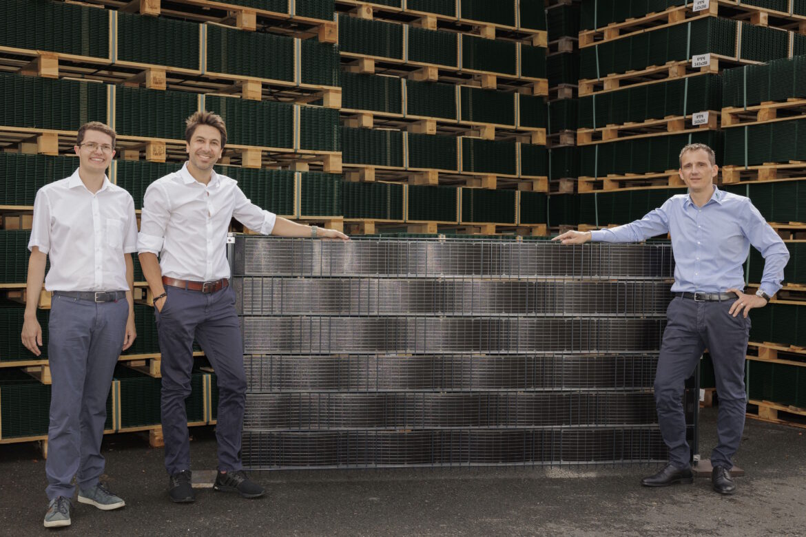 Drei Männer präsentieren den neuen Sunbooster Vertical Solarzaun vor gestapelten Zaunelementen.