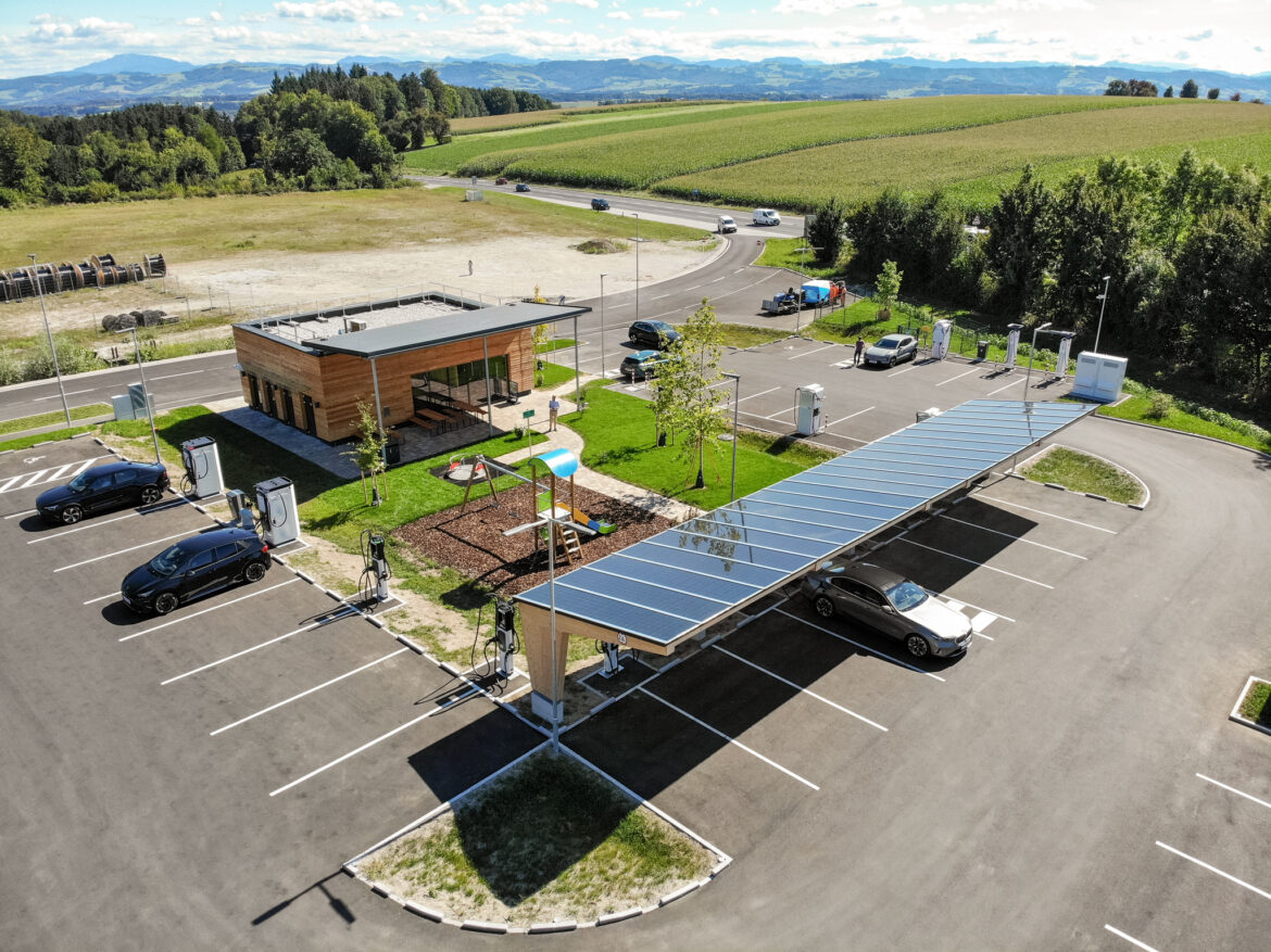 Der moderne Ladepark Amstetten mit Holzgebäude, überdachter Ladeinfrastruktur und angrenzendem Spielplatz inmitten von Feldern.