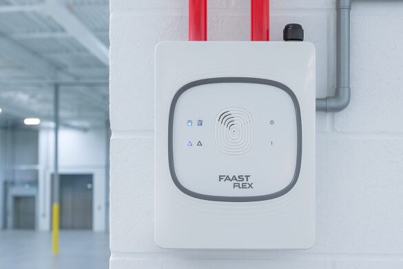 Der Faast Flex Ansaugrauchmelder ist an einer weißen Wand in einer modernen Industriehalle montiert. Zwei rote Ansaugrohre führen in das Gerät, während graue Leitungen seitlich verlaufen. Im Hintergrund sind die helle Hallendecke, Beleuchtung und technische Installationen erkennbar.