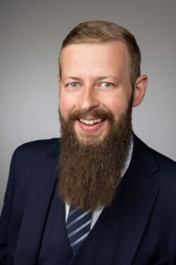 Ein Mann im dunklen Anzug mit langem Vollbart lächelt in einem professionellen Porträt vor neutralem Hintergrund.