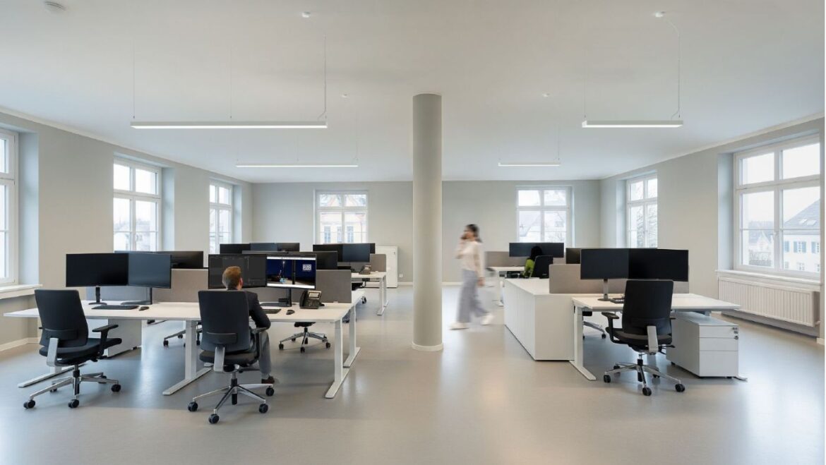 Modernes Großraumbüro mit mehreren Arbeitsplätzen und Pendelleuchten, die für gleichmäßige, blendfreie Beleuchtung sorgen.