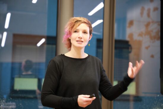 Frau hält einen Presenter in der Hand und erklärt Inhalte während eines Vortrags vor einer Glaswand.