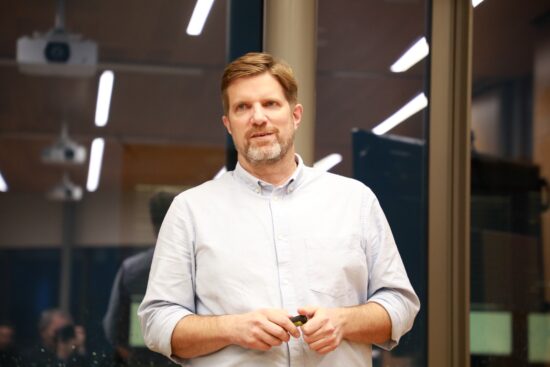 Mann steht vor einer Glaswand und hält einen Presenter in beiden Händen während eines Vortrags.