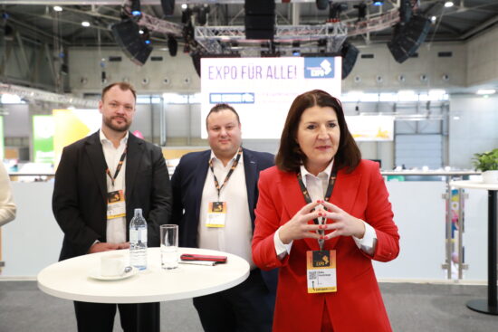 Erika Hochrieser spricht bei der Frauenthal Expo an einem Stehtisch, im Hintergrund stehen zwei weitere Mitglieder des Managements in der Messehalle.