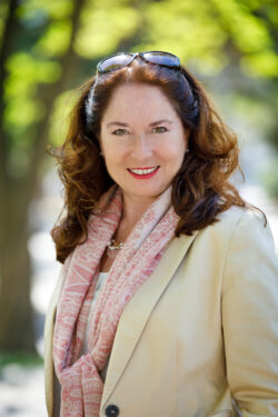 Porträt einer lächelnden Frau im Freien mit Schal und hellem Blazer.