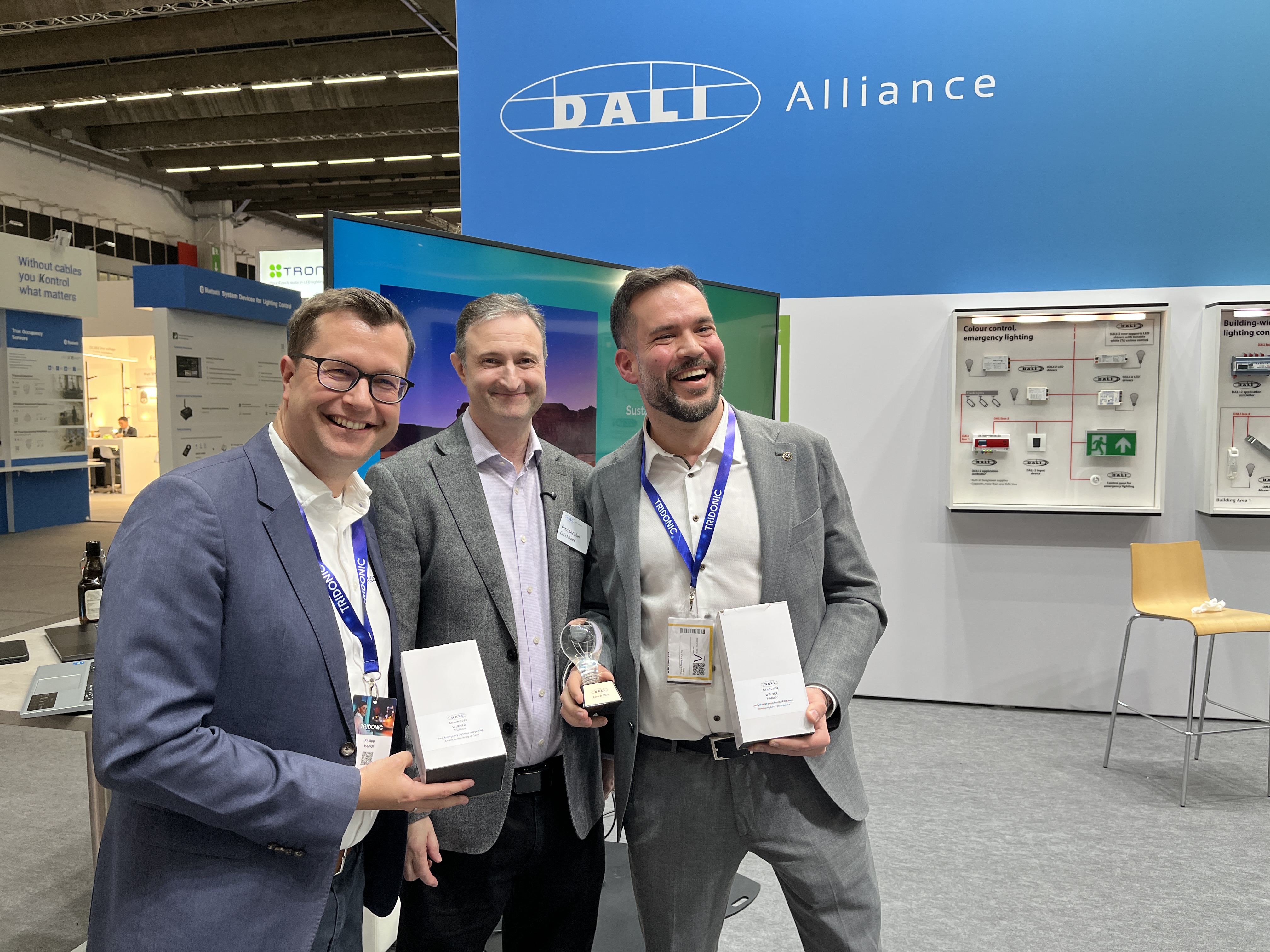 Drei Männer präsentieren Auszeichnungen der Dali Lighting Awards 2026 am Messestand der Dali Alliance.