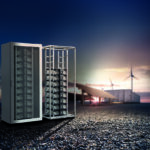 Offener Schaltschrank mit Energiespeichermodulen vor Windrädern und Photovoltaikanlage.