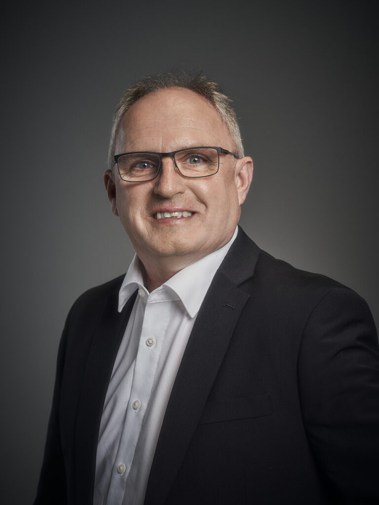 Porträt eines Mannes im Anzug mit Brille vor neutralem Hintergrund.