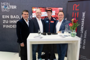 Vier Männer posieren lächelnd nebeneinander hinter einem weißen Stehtisch bei einer Messeveranstaltung. Im Hintergrund ist das Logo „Mein Holter Bad" sowie ein Bildschirm zu sehen.