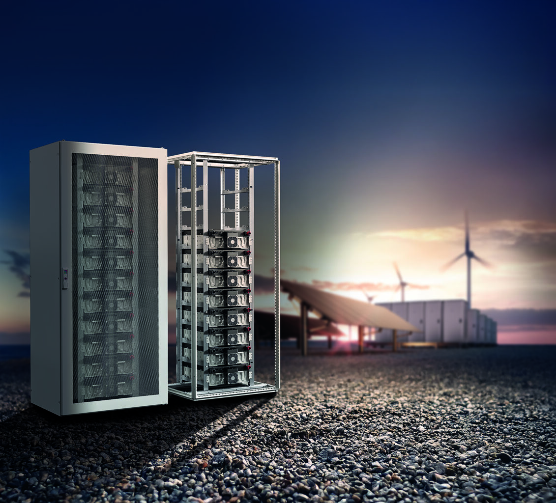 Offener Schaltschrank mit Energiespeichermodulen vor Windrädern und Photovoltaikanlage.