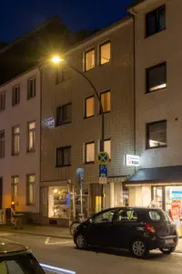 Nächtliche Straßenszene nach der Modernisierung mit gebogener Straßenleuchte vor mehrstöckigen Wohn- und Geschäftshäusern sowie einem parkenden Auto am Fahrbahnrand.