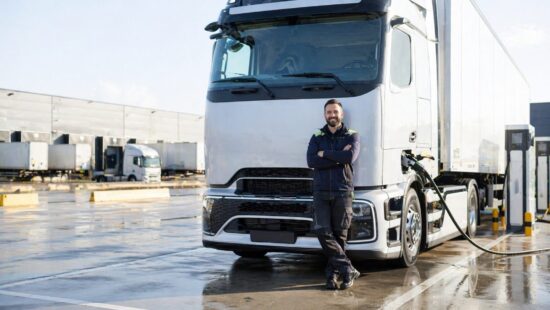 Ein elektrischer schwerer Lkw lädt auf einem modernen Logistikareal, während ein Fahrer in Arbeitskleidung vor dem Fahrzeug steht.