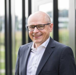 Porträt eines lächelnden Mannes mit Brille im Anzug vor moderner Glasfassade.