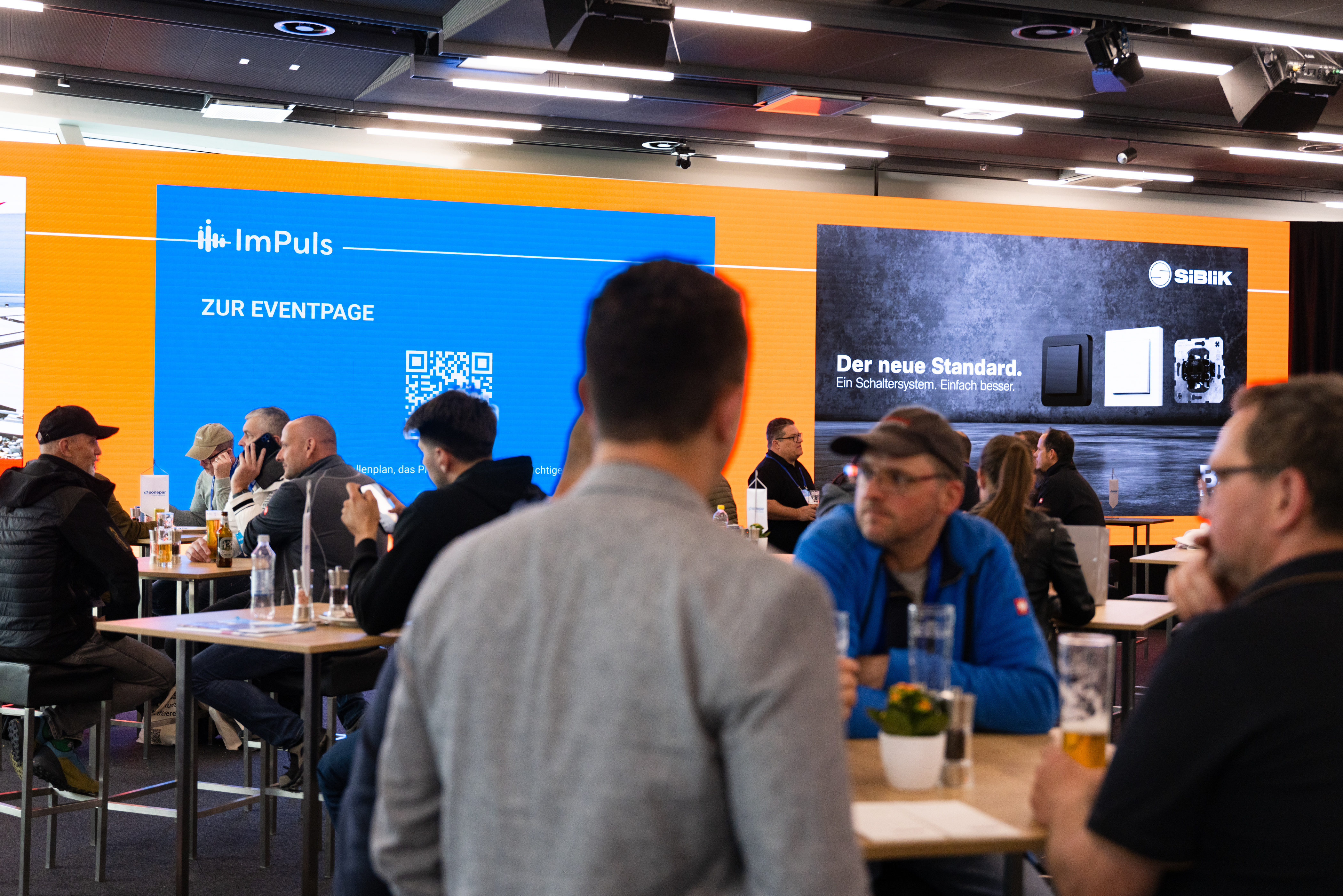 Besucher:innen sitzen im Networkingbereich vor großen digitalen Präsentationsflächen.