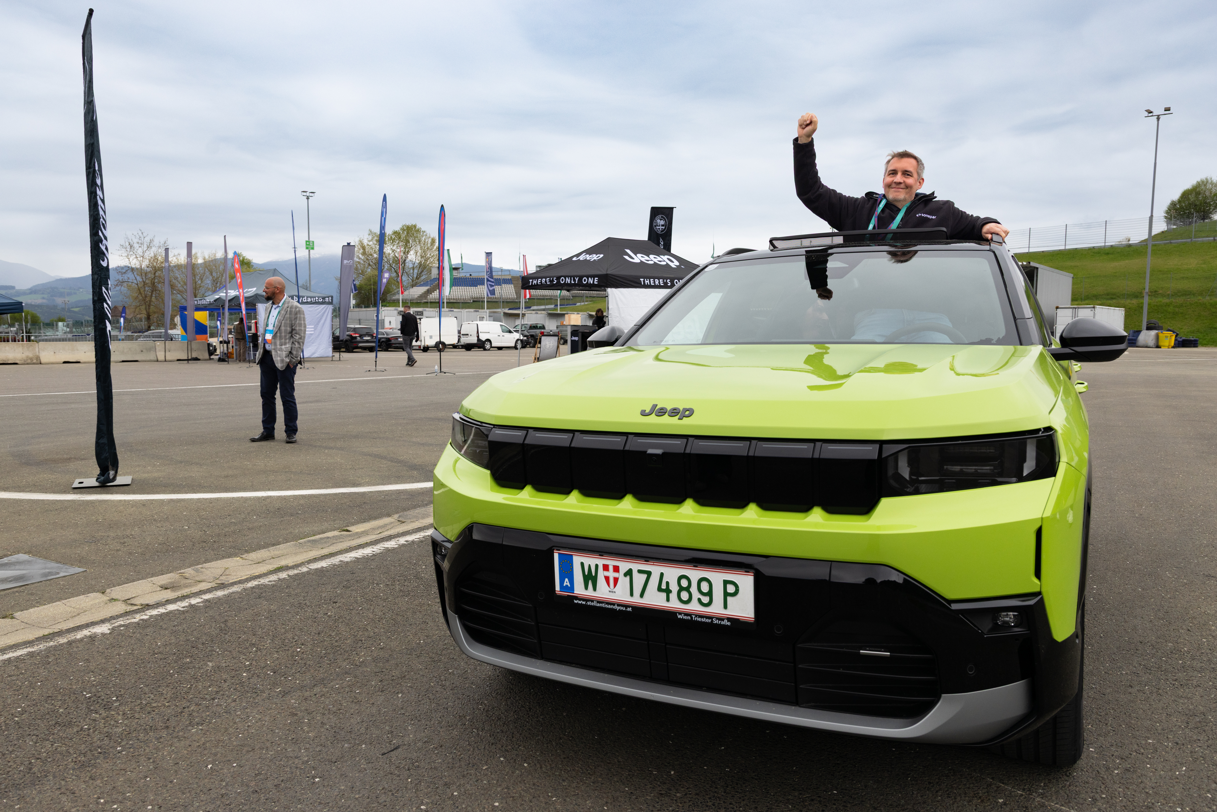 Mann steht jubelnd aus einem grünen Jeep bei einer Outdoor-Veranstaltung auf einer Rennstrecke