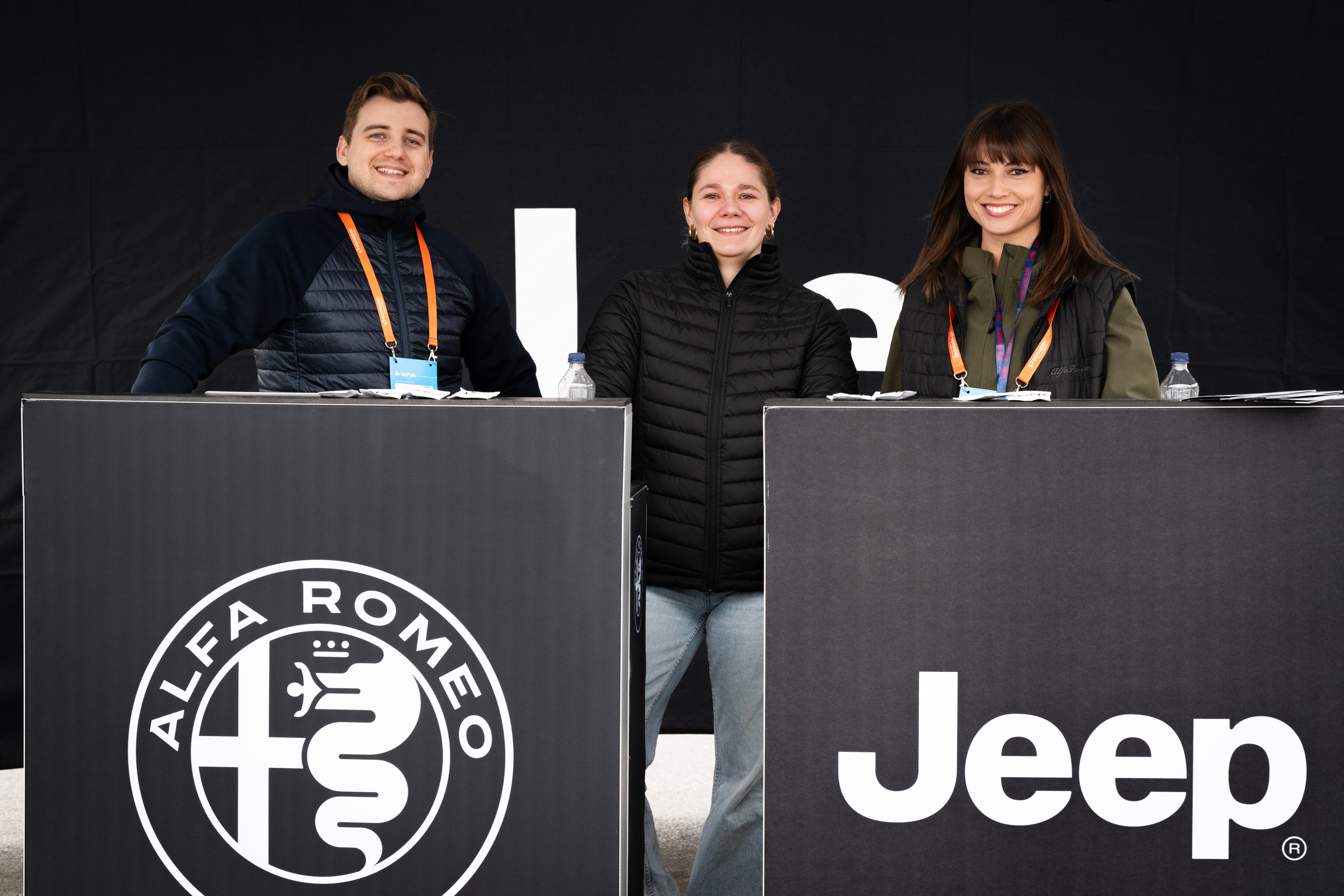 Drei Mitarbeitende stehen hinter Empfangstheken mit Alfa Romeo- und Jeep-Logos.