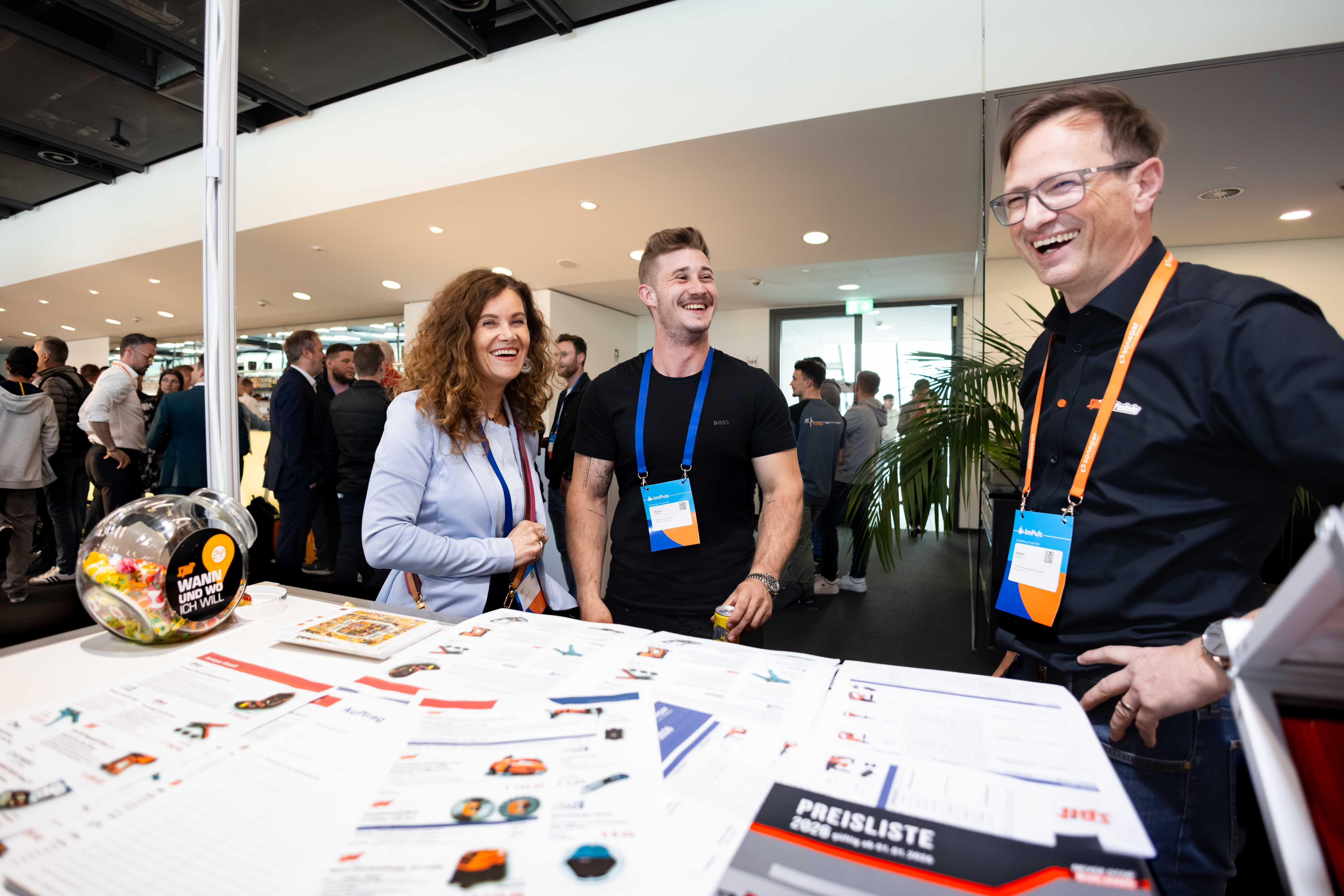 Drei Personen lachen gemeinsam an einem Messestand mit Produktunterlagen.