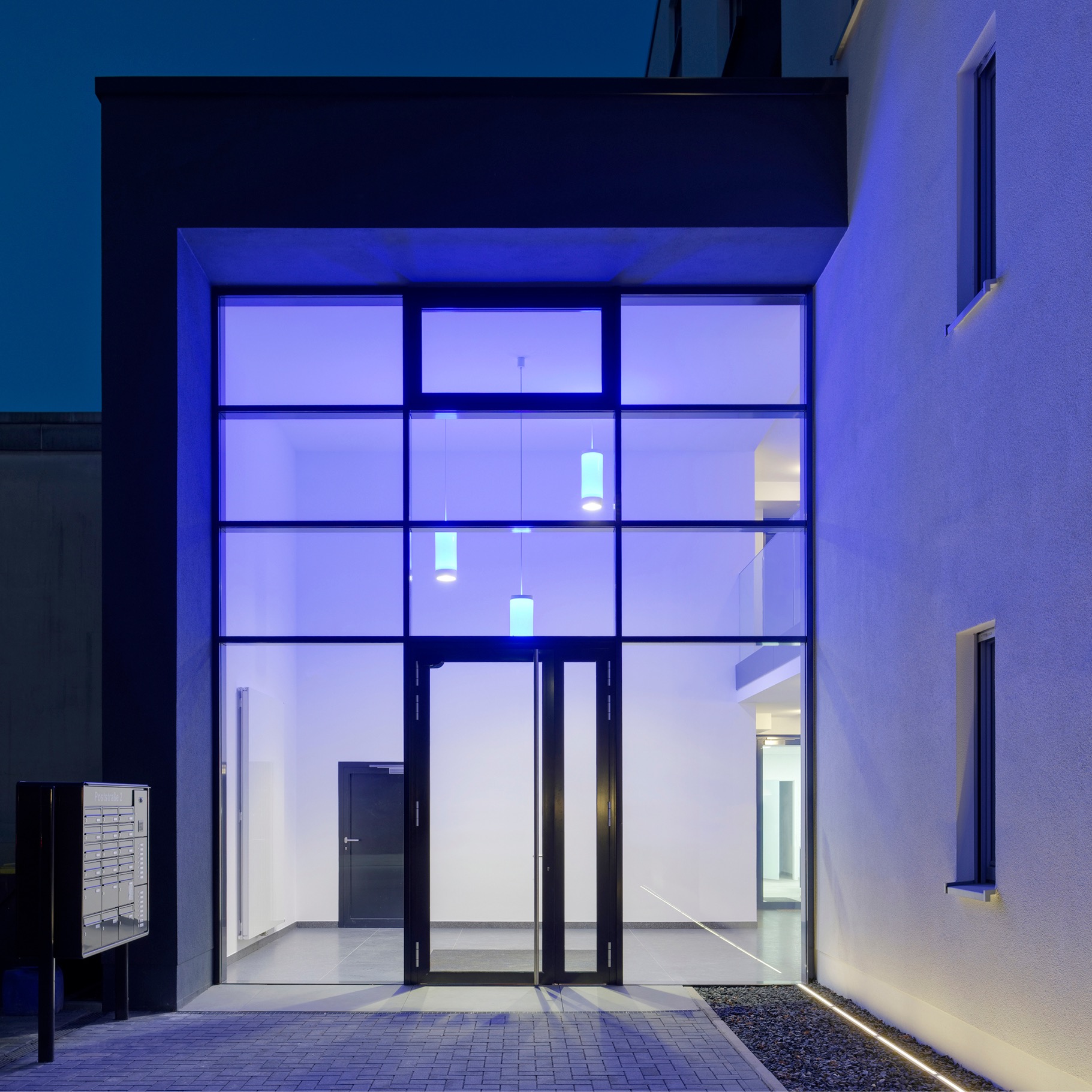Moderner Gebäudeeingang mit großer Glasfassade, Innenbeleuchtung und blauer Lichtstimmung bei Nacht.