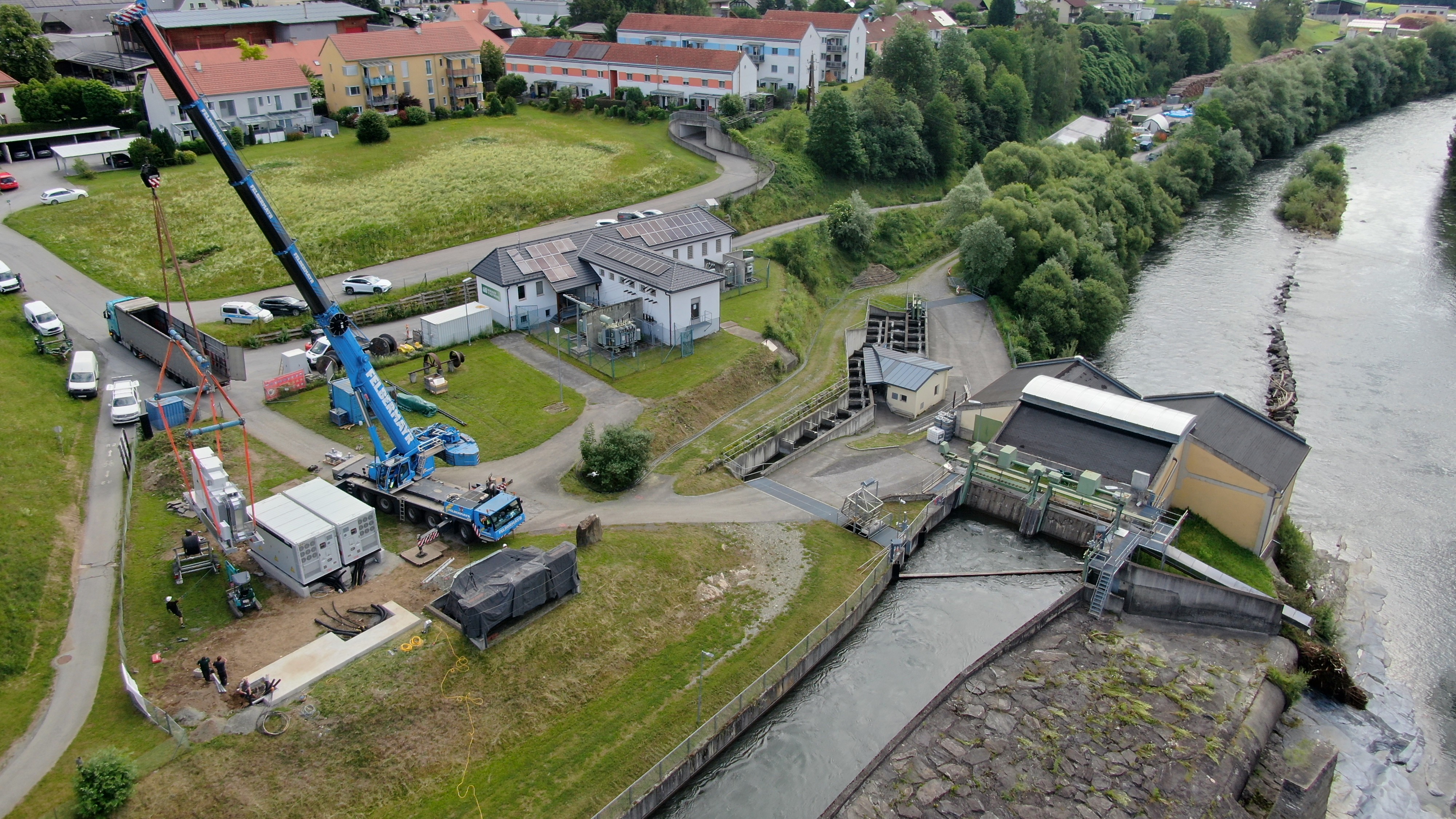 Luftaufnahme eines Wasserkraftwerks mit Baustelle, Kran und Batteriespeicher zur Absicherung der regionalen Energieversorgung.