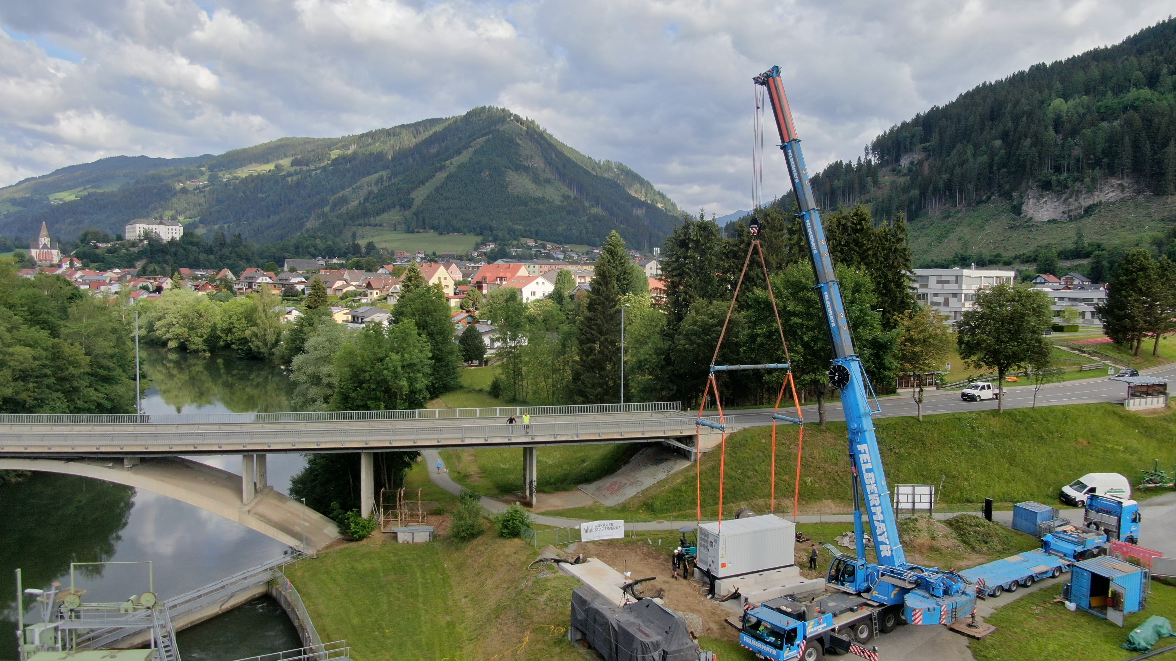 Kran hebt einen Batteriespeicher in einer alpinen Umgebung nahe Brücke, Fluss und Wasserkraftwerk.
