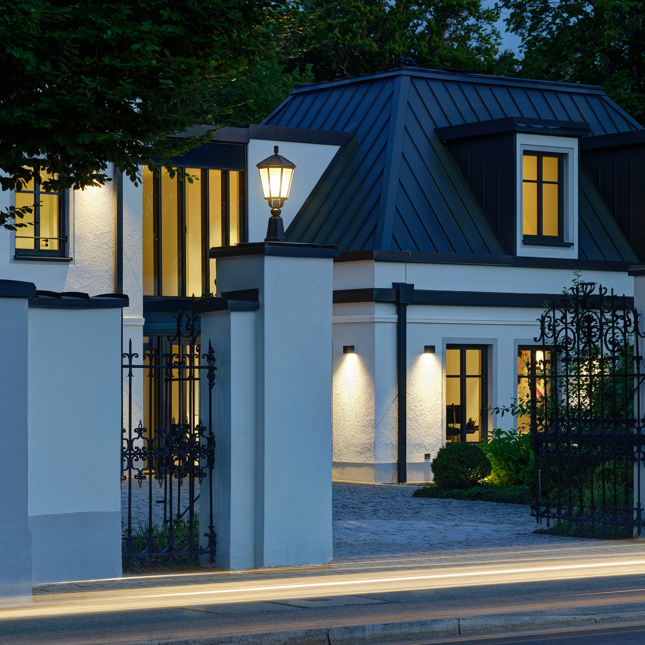Villa mit beleuchteter Fassade, Gartenleuchten, Eingangstor und warmem Innenlicht bei Nacht.