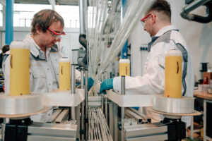 Zu sehen ist ein weitläufiges Solarfeld in einer ländlichen Landschaft. Mehrere Reihen von Photovoltaikmodulen erstrecken sich über eine grüne Wiese mit Blumen und Gräsern. Die Module sind auf geneigten Tragkonstruktionen montiert, die für Stabilität und optimale Sonneneinstrahlung sorgen. Im Hintergrund befinden sich Felder, Bäume und ein bewaldeter Höhenzug unter einem teilweise bewölkten Himmel. Das Bild verdeutlicht den Einsatz moderner Leichtbau- und Materialtechnologien für erneuerbare Energien.