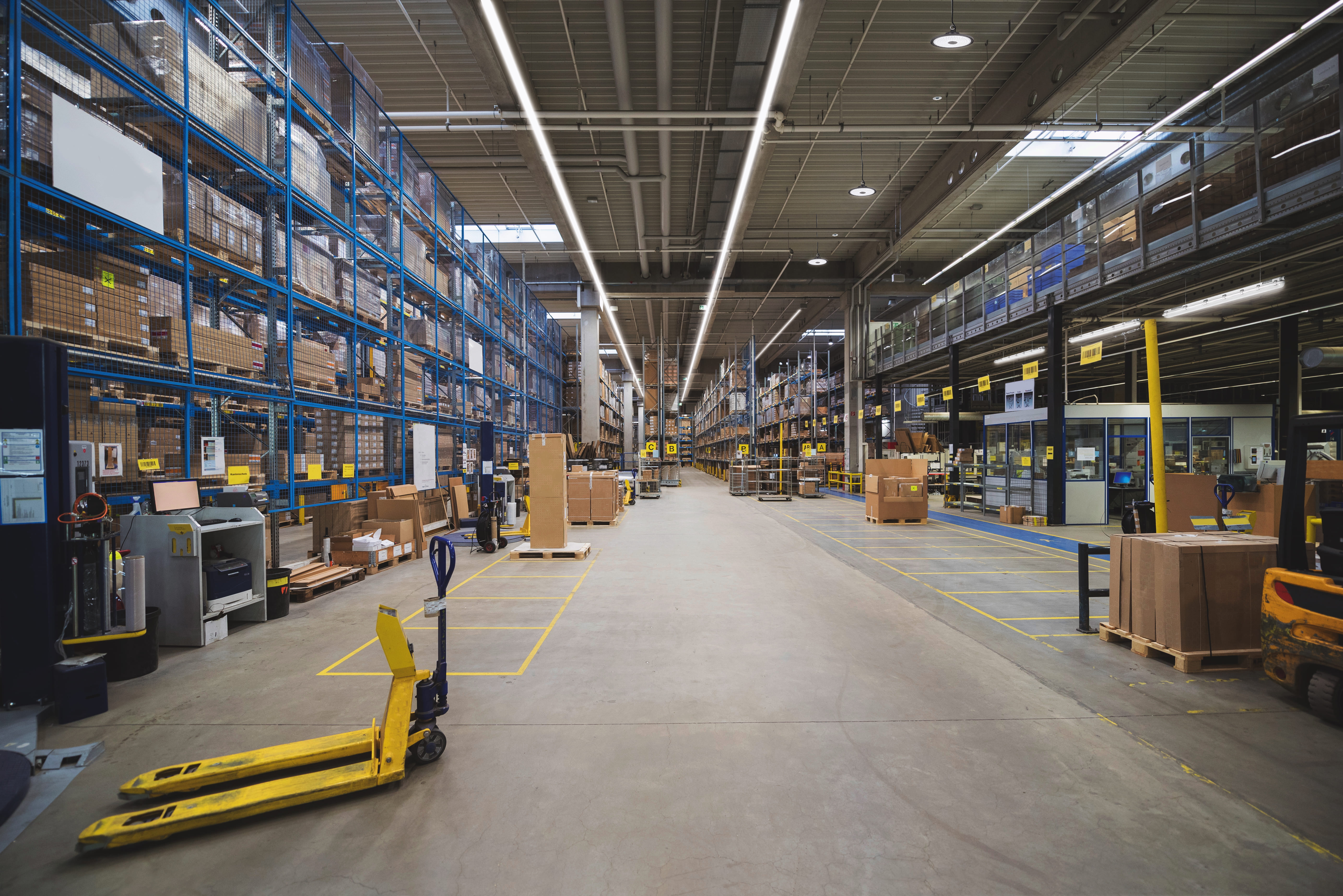 Große Logistikhalle mit Hochregallagern, Arbeitsbereichen und moderner LED-Beleuchtung.